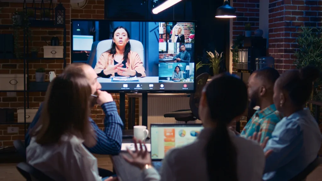Five people in a modern office around a table in a video conference with remote participants on a wall-mounted screen
